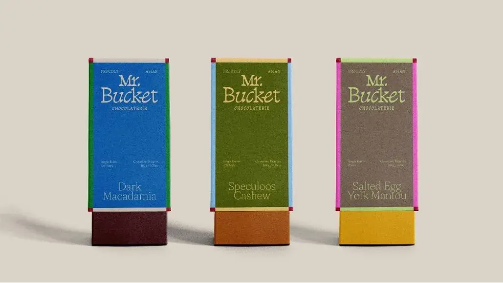 Three chocolate bars with colorful packaging labeled: Dark Macadamia, Speculoos Cashew, and Salted Egg Yolk Mantou by Mr. Bucket Chocolaterie.