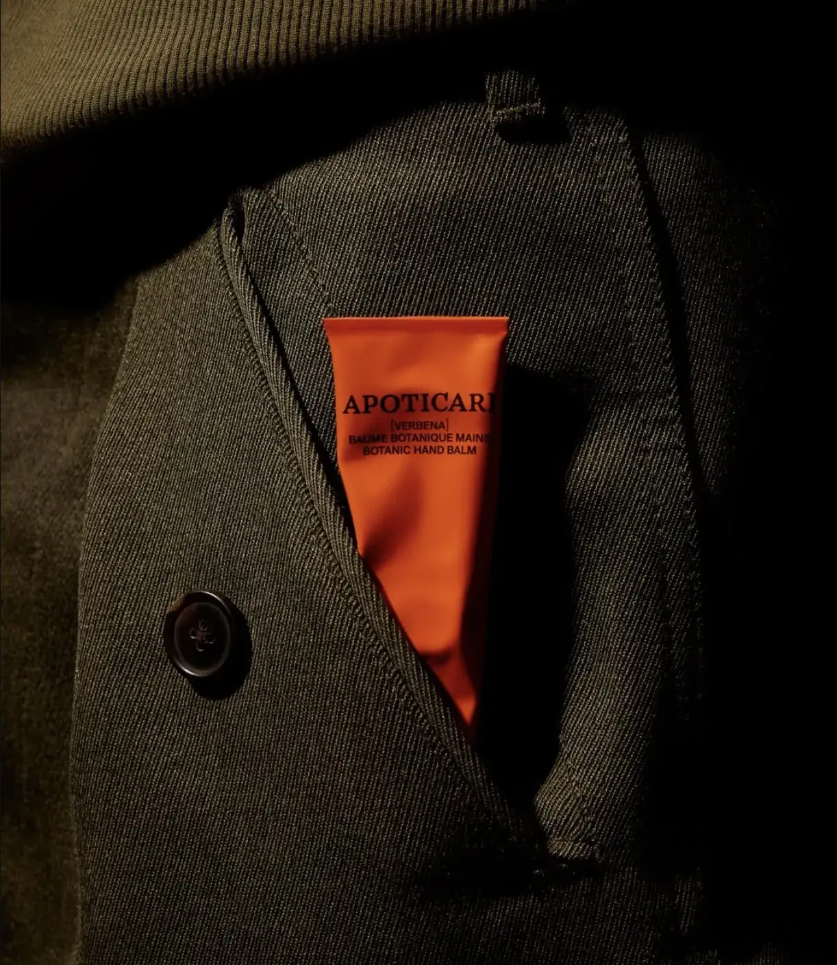 A tube of Apoticare hand balm partially visible in the pocket of a dark coat, illuminated by warm light.