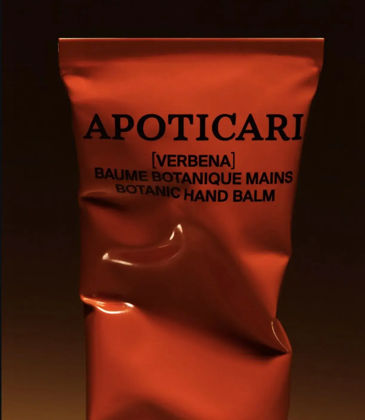Orange tube of Apoticari Verbena Botanic Hand Balm against a dark background, displaying bold black text and a shiny, crinkled surface.