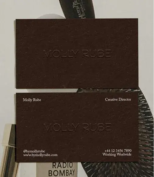 Two brown business cards for Molly Rube, Creative Director, featuring embossed text and contact details. Background includes a brush and books.