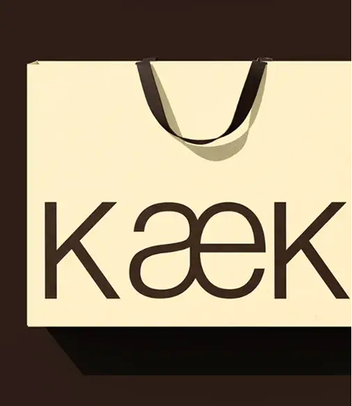 A cream shopping bag with a black handle and the text “kæek” printed in bold brown letters, set against a dark background.