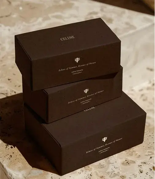 Three stacked brown boxes on a textured surface, labeled "Celine" with decorative logos and text "Essence of Summer, Essence of Nature."