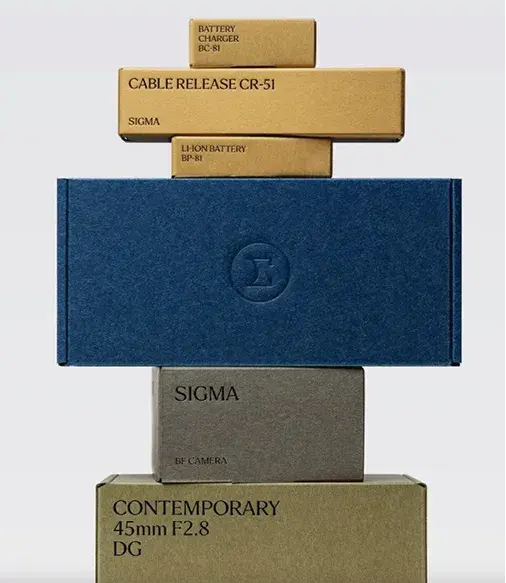 Stacked product boxes of various sizes, including a blue box with a logo, labeled Sigma, items like a cable release, battery, and camera lens.